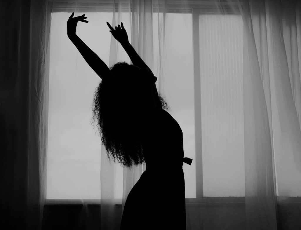 A woman with long curly hair is dancing with her arms raised above her in front of a window with curtains.